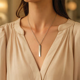 A close-up of a woman wearing a silver bar pendant necklace with engraved text, styled with a beige blouse.