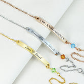 Four personalized bar bracelets arranged diagonally on a white textured fabric background. The bracelets are in gold, silver, and rose gold tones, each featuring an engraved nameplate with Roman numerals or sentimental text ('Forever in my heart', 'Liam x Sarah'), a small faceted crystal bead, and an adjustable chain.