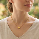 A close-up lifestyle shot of a woman wearing a delicate gold heart pendant necklace on a fine chain, paired with a light beige linen top.