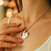 A close-up lifestyle shot of a woman wearing a silver heart-shaped pendant necklace with a white crescent moon and star design, set against a warm, softly blurred background.