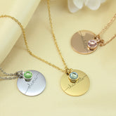 Three stainless steel engraved name necklaces with round pendants featuring different birthstones (emerald, pink, and amethyst) and personalized engraved names (Eloise, Hannah, and Thomas) on a gold, silver, and rose gold chain, displayed against a soft-focus background.