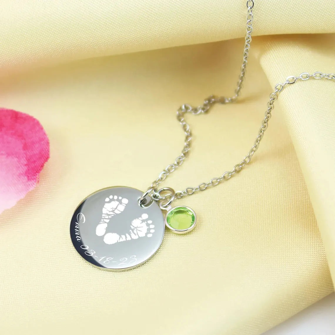 A stainless steel necklace with a footprint-shaped pendant and a small green Swarovski crystal charm, displayed on a yellow fabric background with a hint of pink.