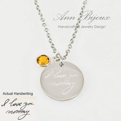 "The pendant includes a name and date etched along the bottom edge, paired with a small amber-colored birthstone charm on a delicate chain."