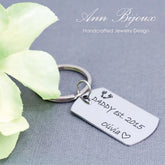A stainless steel key chain with a laser engraved inscription 'Daddy Est' alongside a small footprint design, personalized with a name and date.