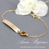 A gold-filled bracelet with a flat cable chain and a central Swarovski birthstone, displayed on a grey background with a white flower and a name tag.