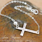 Silver bracelet with a cross pendant and pearl on a stone surface, branded 'Ann Bijoux'.