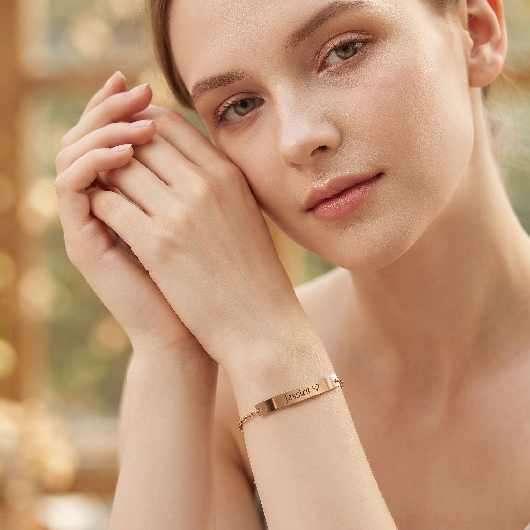 A close-up lifestyle shot of a woman wearing a delicate rose gold bar bracelet engraved with 'HELLO' and a small heart, set against a warm, softly blurred bokeh background.