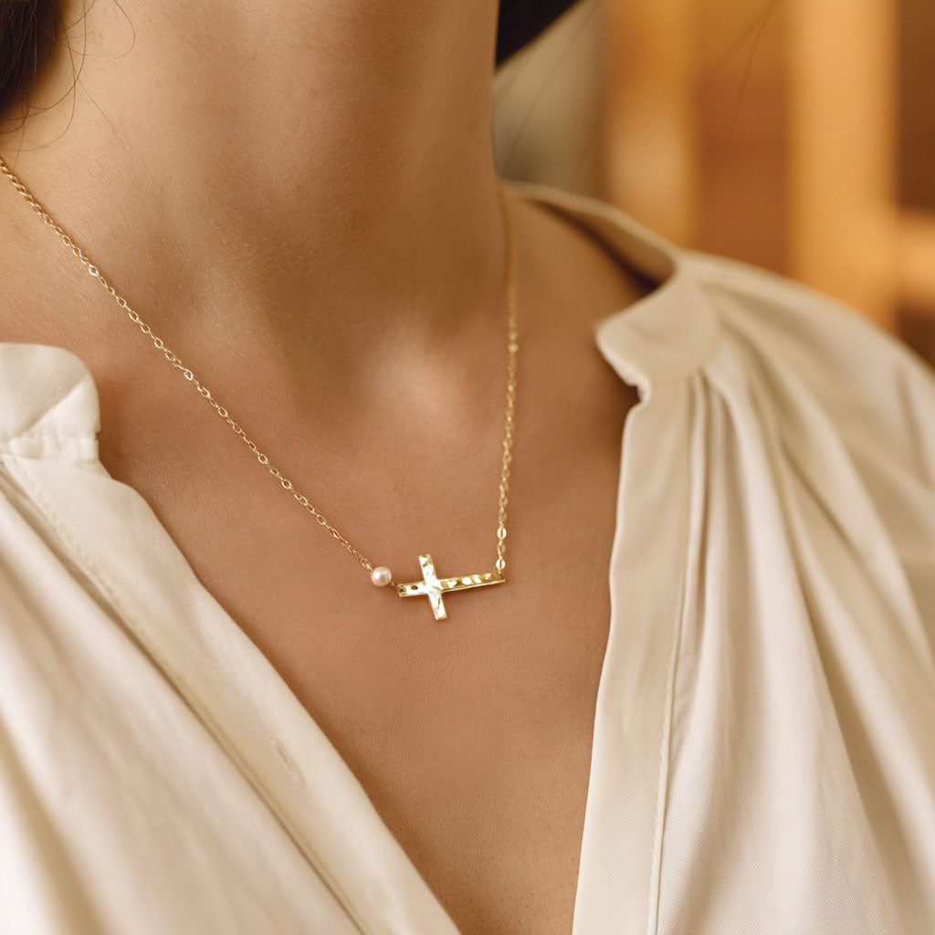 A close-up lifestyle shot of a model wearing a delicate gold necklace with a small cross pendant, a tiny pearl bead, and a tiny clear stone, styled with a cream-colored blouse.