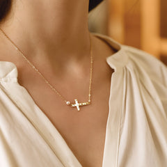 A close-up lifestyle shot of a model wearing a delicate gold necklace with a small cross pendant, a tiny pearl bead, and a tiny clear stone, styled with a cream-colored blouse.