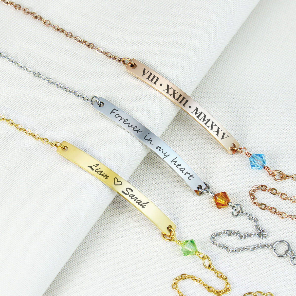 Four personalized bar bracelets arranged diagonally on a white textured fabric background. The bracelets are in gold, silver, and rose gold tones, each featuring an engraved nameplate with Roman numerals or sentimental text ('Forever in my heart', 'Liam x Sarah'), a small faceted crystal bead, and an adjustable chain.