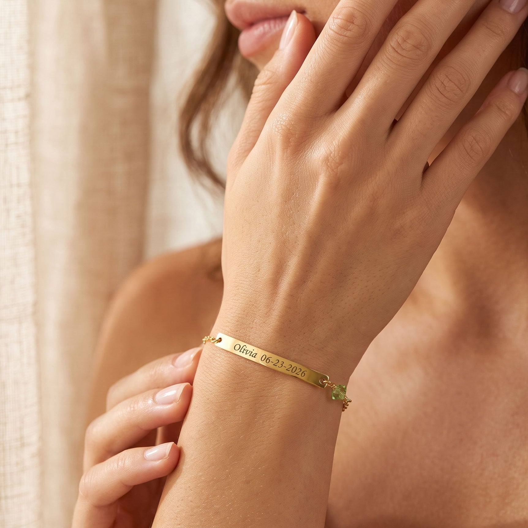 A close-up of a woman's wrist wearing a slim gold bracelet with engraved text and a small green gemstone accent, set against a soft beige background.