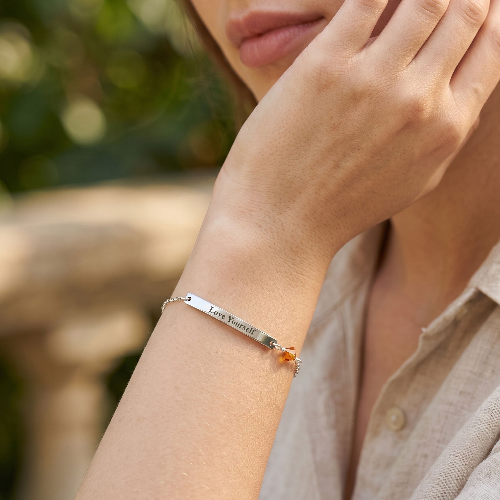 A close-up lifestyle shot of a woman wearing a silver bar bracelet engraved with 'Love Yourself' on her wrist, paired with a light beige linen shirt against a soft, blurred outdoor background.