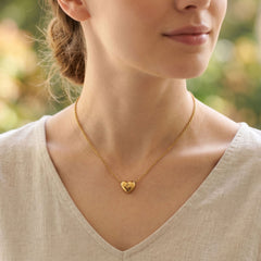 A close-up lifestyle shot of a woman wearing a delicate gold heart pendant necklace on a fine chain, paired with a light beige linen top.