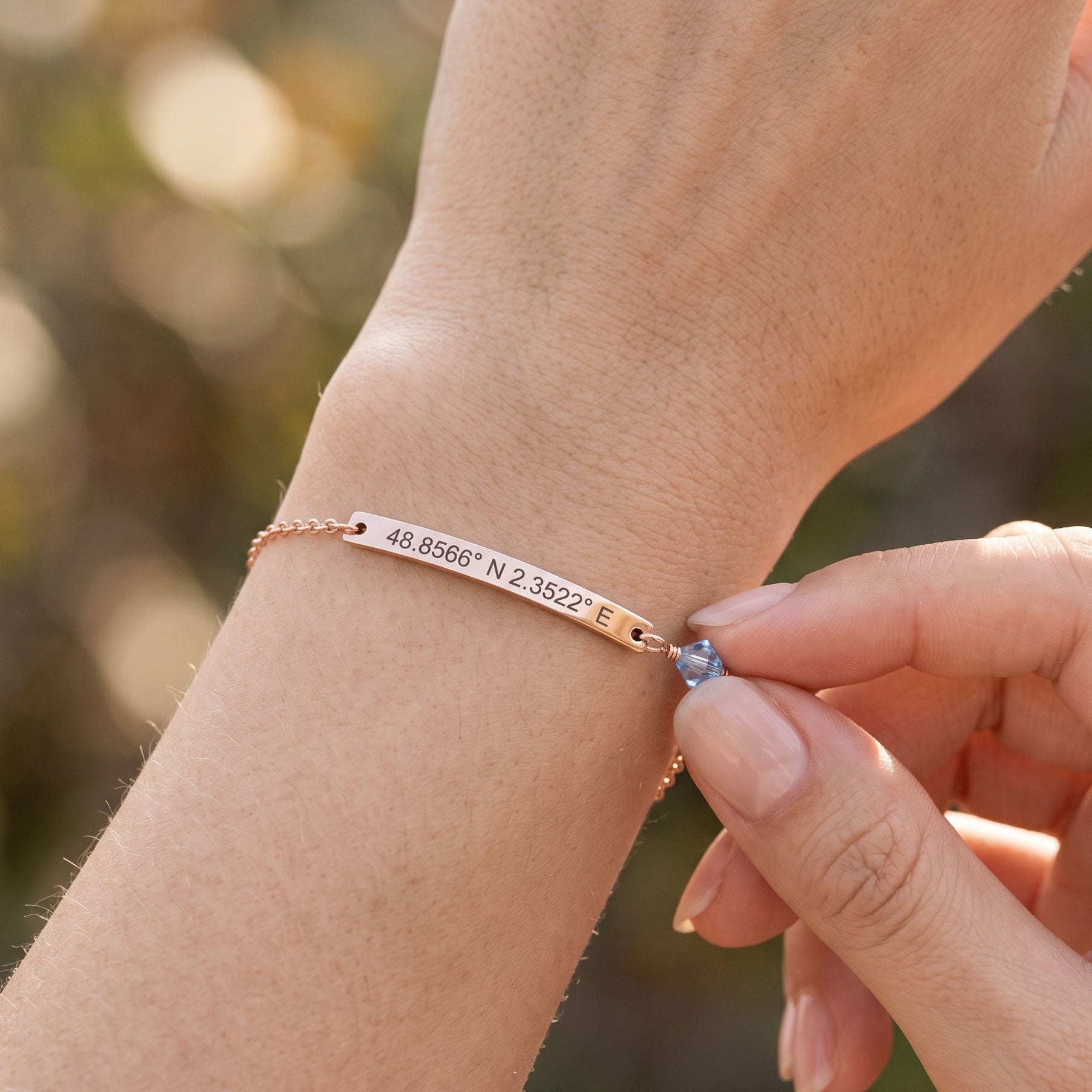 A close-up lifestyle shot of a person's wrist wearing a rose gold chain bracelet with a rectangular engraved plate showing coordinates, with another hand adjusting a small blue gemstone charm.