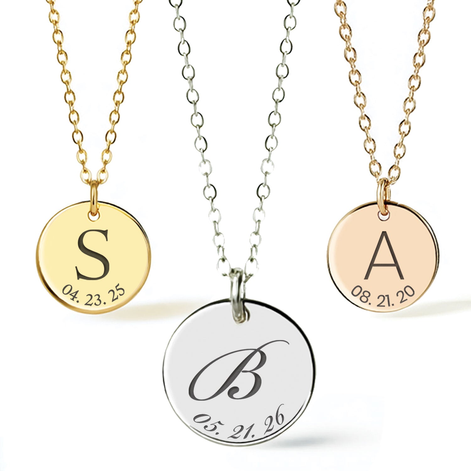 Three personalized round pendant necklaces on chains, shown against a clean white background. The left necklace has a gold chain and a gold pendant engraved with the letter S and a date.