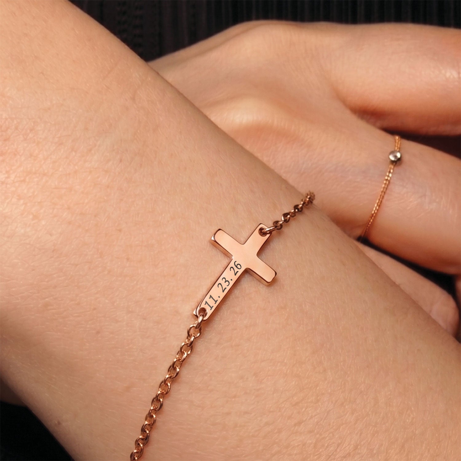 Rose gold cross bracelet with engraved date on wrist against a dark background