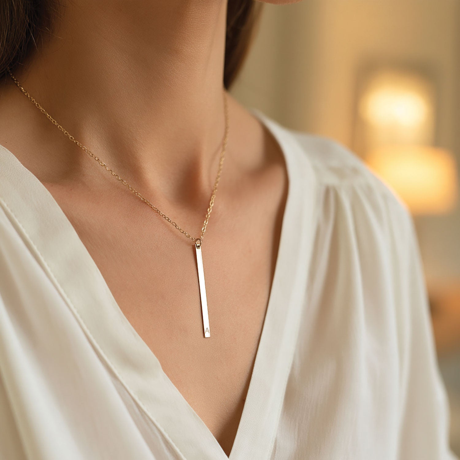 A close-up of a woman wearing a delicate gold chain necklace with a slim vertical bar pendant, styled with a white blouse against a softly blurred warm indoor background.