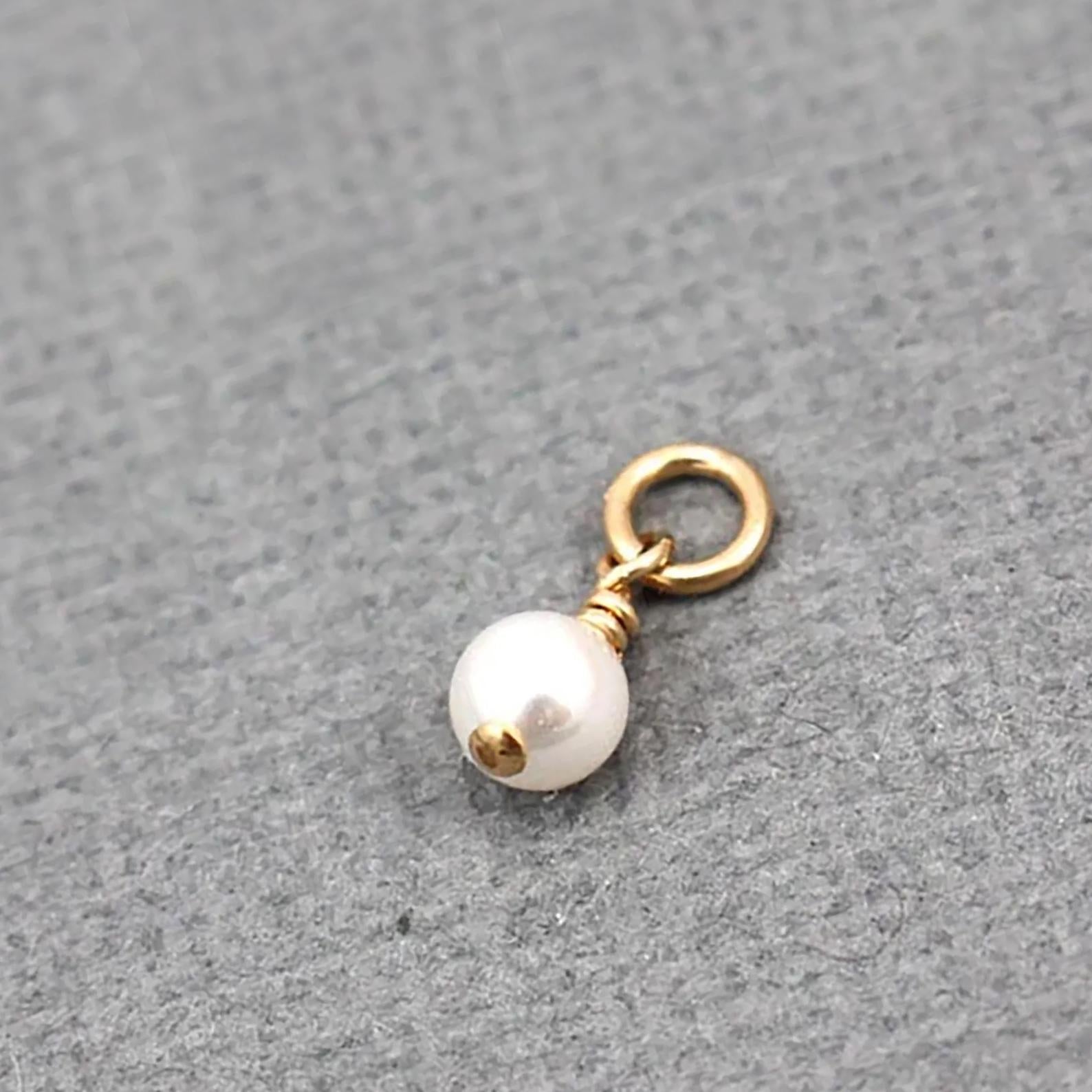 A small jewelry charm featuring a single white pearl bead with a gold cap and a gold jump ring, displayed on a textured gray background.