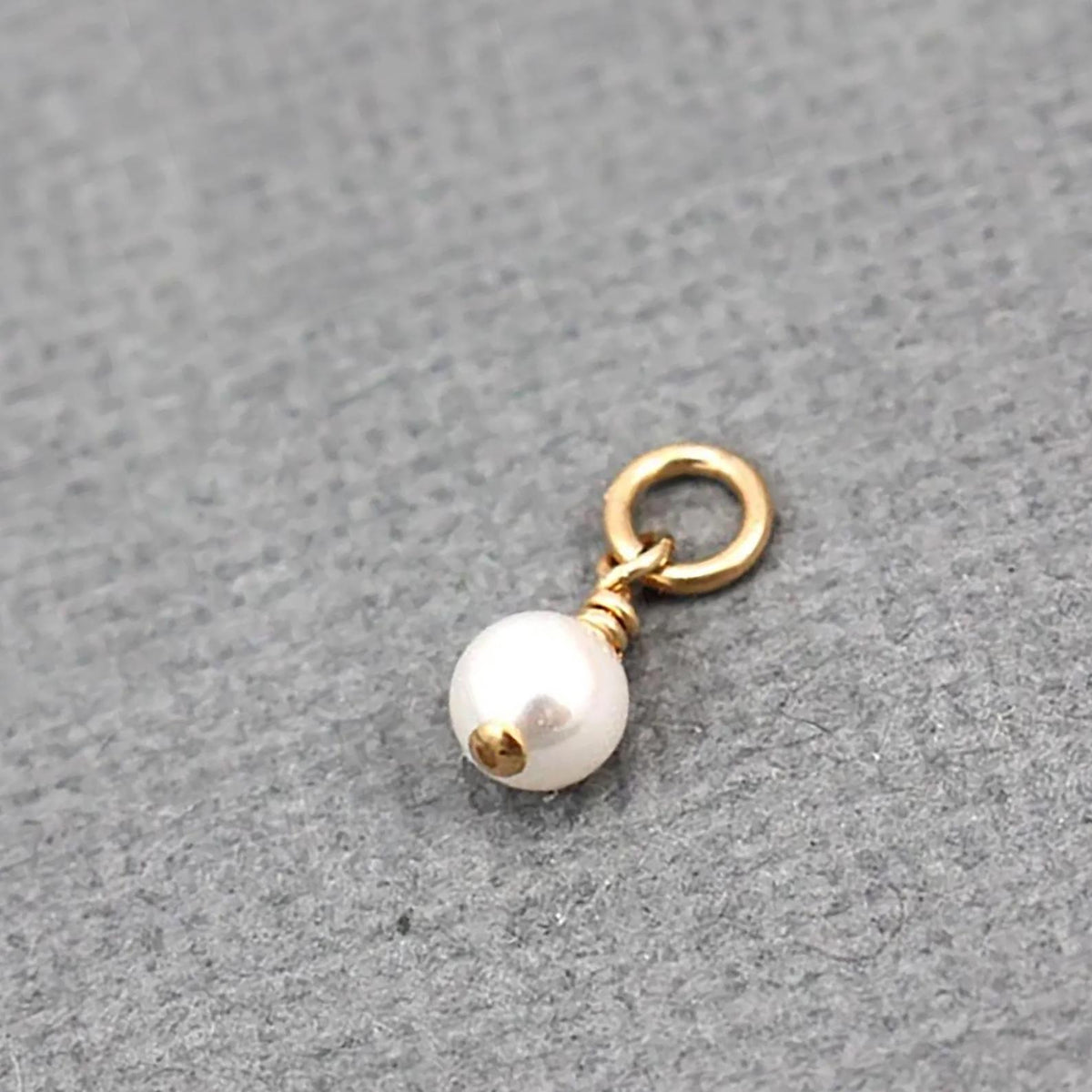 A small jewelry charm featuring a single white pearl bead with a gold cap and a gold jump ring, displayed on a textured gray background.
