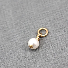 A small jewelry charm featuring a single white pearl bead with a gold cap and a gold jump ring, displayed on a textured gray background.