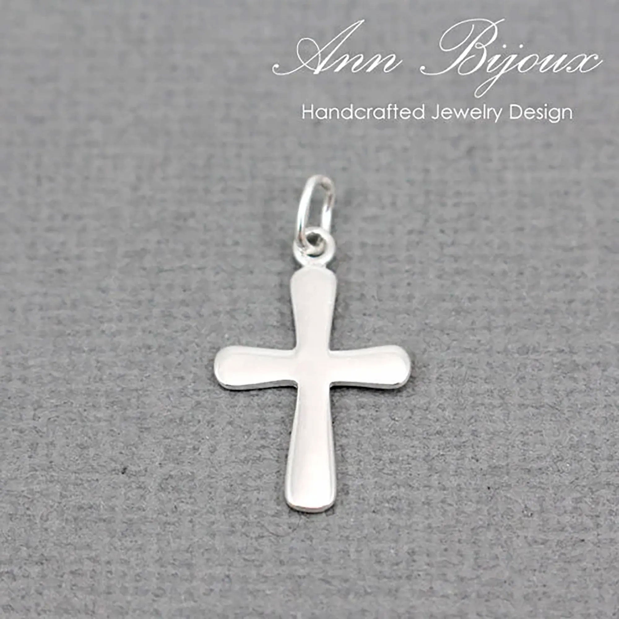 A polished silver cross pendant with a smooth, flat surface and a small top loop, displayed on a textured gray background with 'Ann Bijoux Handcrafted Jewelry Design' written in white script at the top.