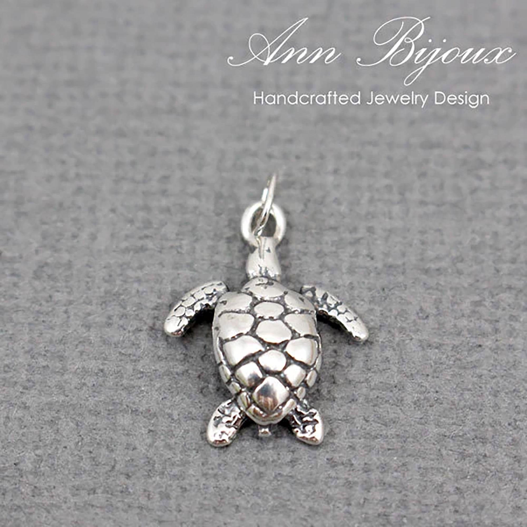 A silver sea turtle pendant charm with a textured shell and outstretched flippers, shown on a gray textured background with the brand name 'Ann Bijoux' at the top.