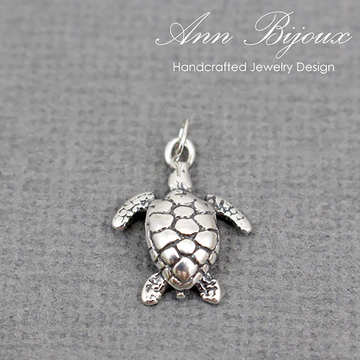 A silver sea turtle pendant charm with a textured shell and outstretched flippers, shown on a gray textured background with the brand name 'Ann Bijoux' at the top.