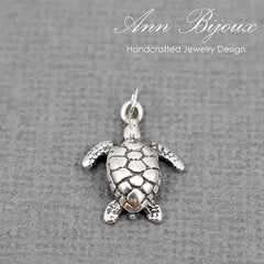 A silver sea turtle pendant charm with a textured shell and outstretched flippers, shown on a gray textured background with the brand name 'Ann Bijoux' at the top.