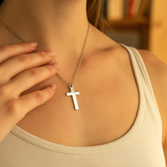 A close-up of a person wearing a silver cross pendant necklace on a delicate chain, paired with a beige ribbed tank top. The background is a warm, softly blurred indoor setting.