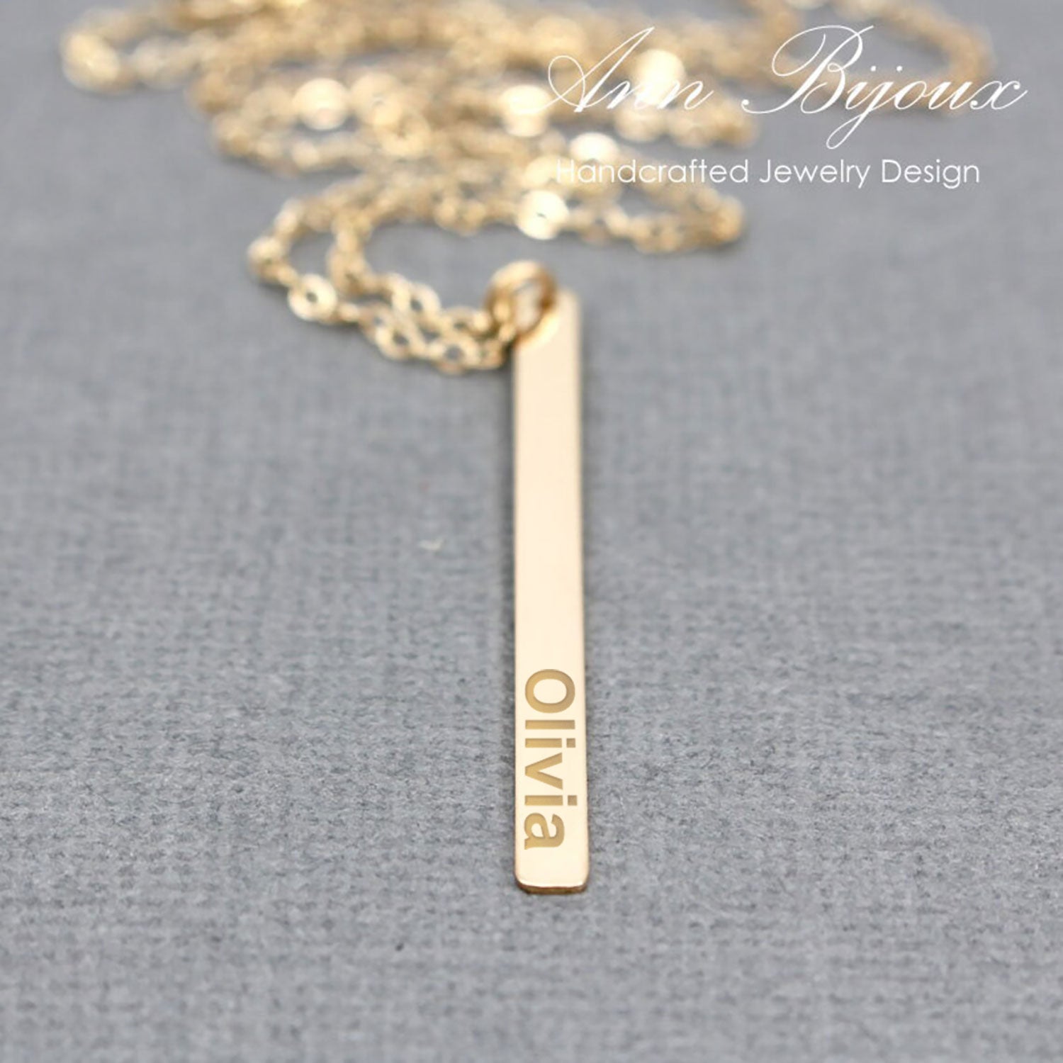 A gold bar necklace with the name 'Olivia' engraved vertically, displayed on a textured gray background with a gold chain partially visible at the top. The brand name 'Ann Bijoux Handcrafted Jewelry Design' is watermarked in the upper right.