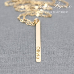 A gold bar necklace with the name 'Olivia' engraved vertically, displayed on a textured gray background with a gold chain partially visible at the top. The brand name 'Ann Bijoux Handcrafted Jewelry Design' is watermarked in the upper right.