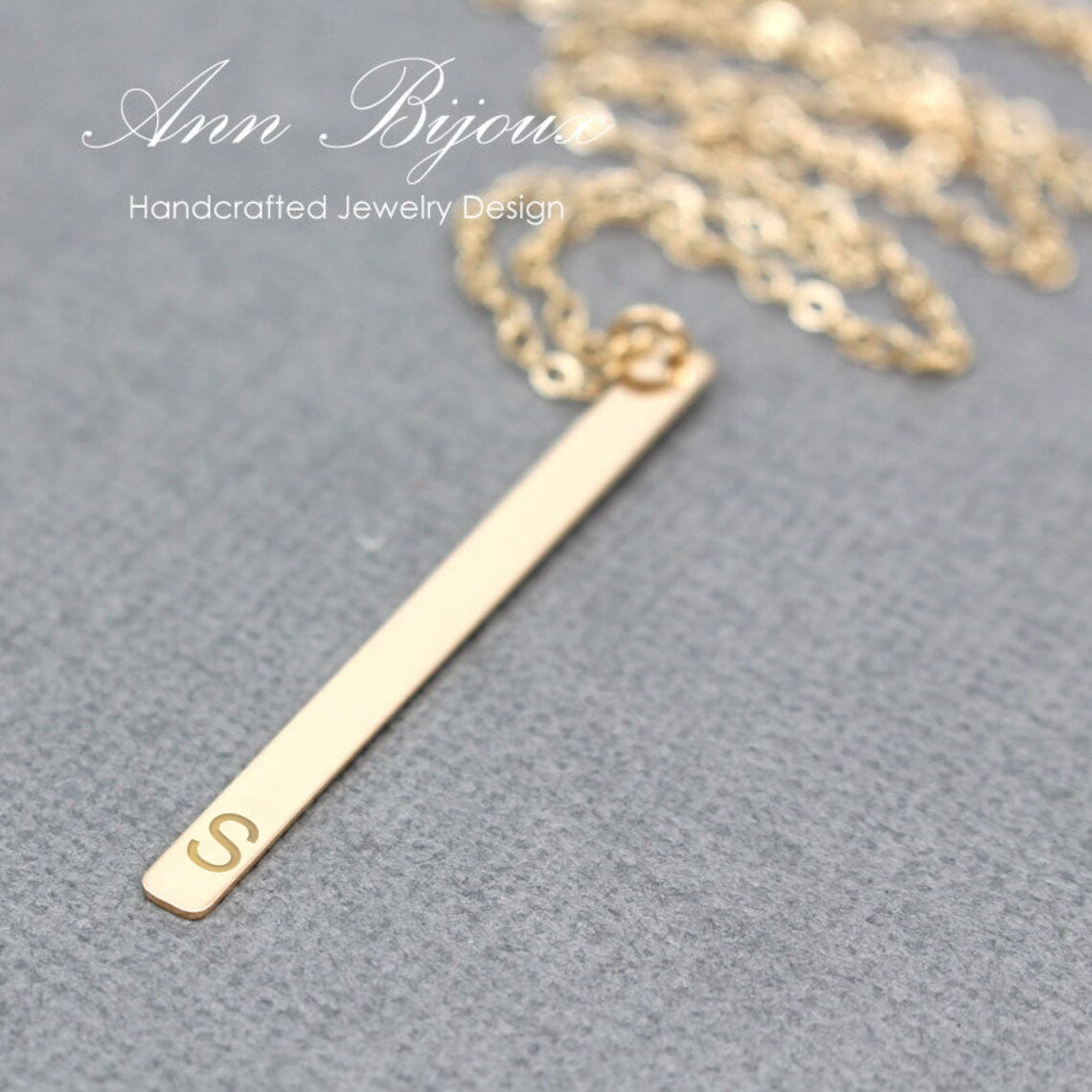 A gold bar pendant necklace with a small engraved letter 'S' at one end, resting on a textured gray surface with a gold chain blurred in the background. White text overlay reads 'Ann Bijoux Handcrafted Jewelry Design'.