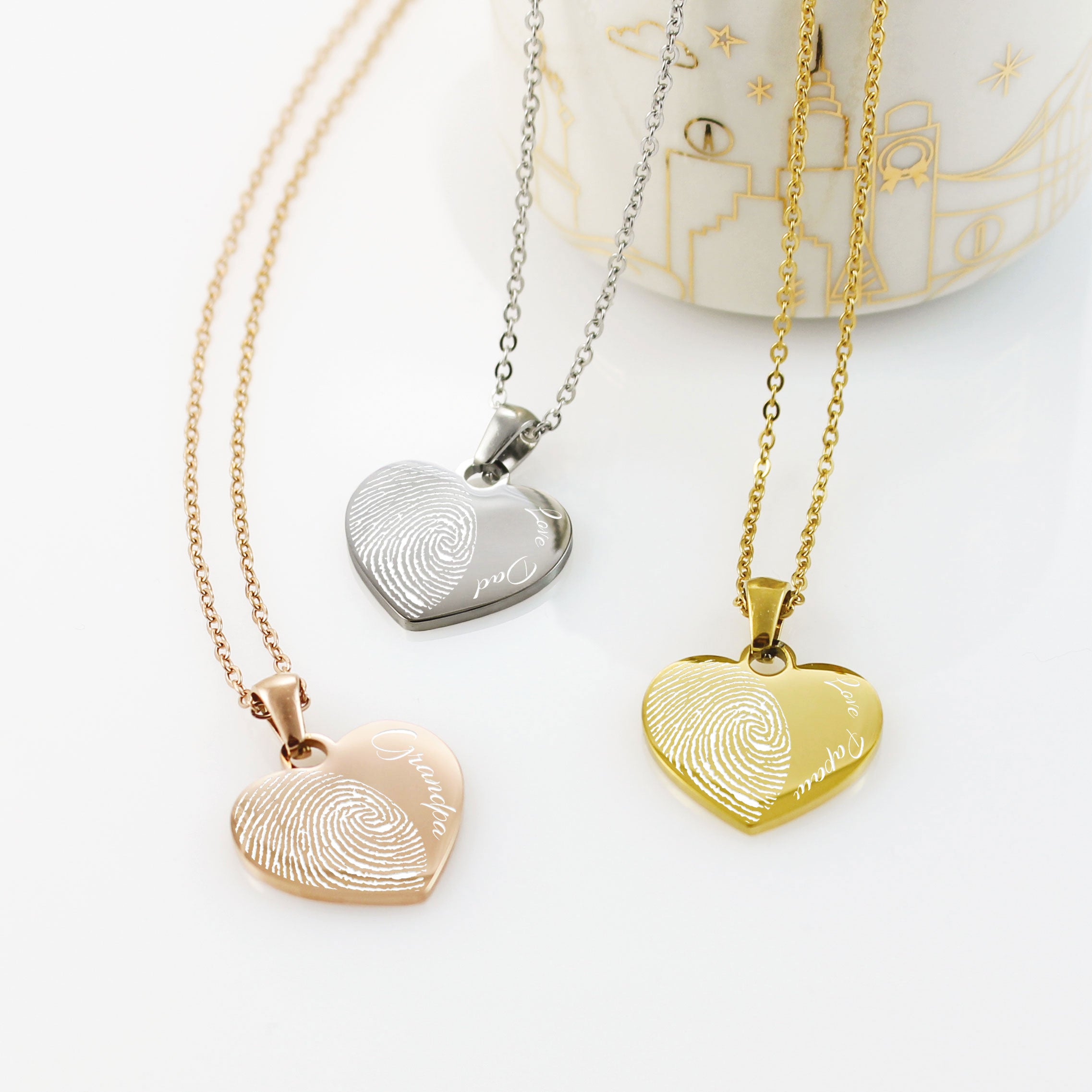 "Three custom heart-shaped fingerprint necklaces in rose gold, silver, and yellow gold. Each pendant features a detailed half-heart fingerprint engraving paired with personalized names like 'Grandpa,' 'Love Dad,' and 'Love Papa' in an elegant script font."