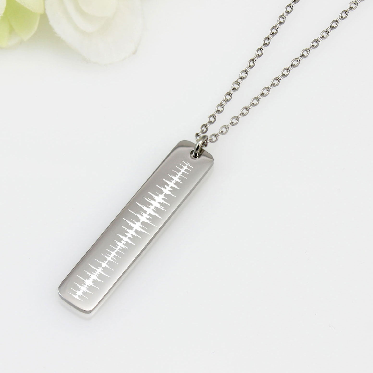 A polished silver rectangular bar pendant hanging vertically from a silver chain, engraved with a detailed white sound wave pattern against a white background with a floral accent.