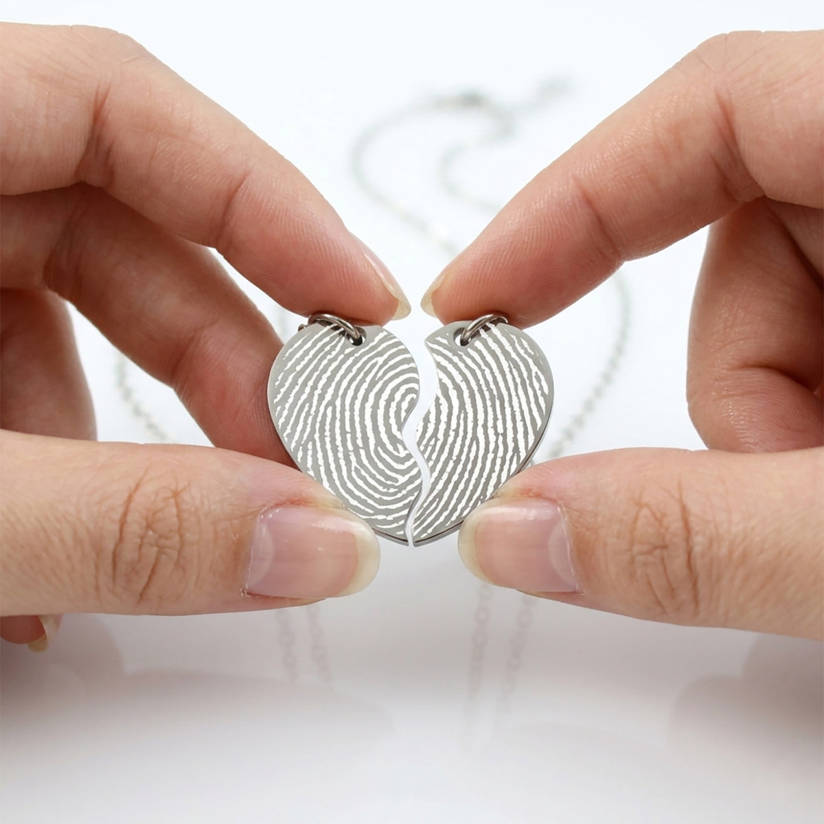 Two hands holding a silver heart-shaped fingerprint pendant against a white background