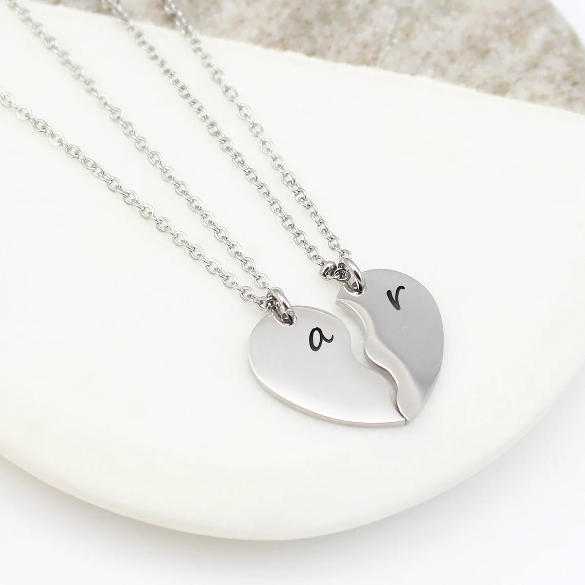 Two silver heart-half necklaces that fit together to form a full heart. Each piece is engraved with a single initial—'S' and 'K'—in a delicate black script font, hanging from matching cable chains.