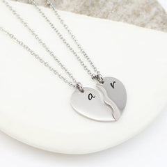 Two silver heart-half necklaces that fit together to form a full heart. Each piece is engraved with a single initial—'S' and 'K'—in a delicate black script font, hanging from matching cable chains.