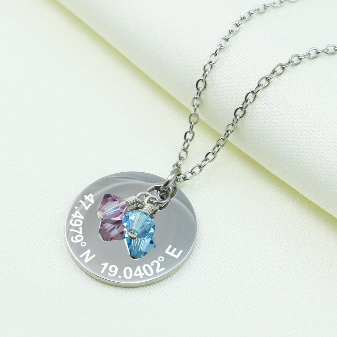 Personalized silver coordinates necklace featuring a round disc engraved with GPS locations, accented by purple and light blue crystal charms.