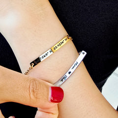 Two bracelets on a wrist with engraved text, one gold and one silver.