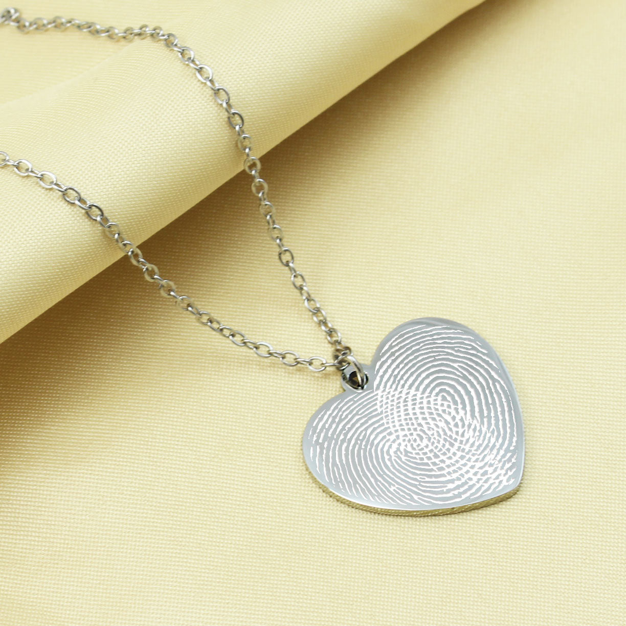 Silver heart-shaped fingerprint pendant on a chain against a beige background