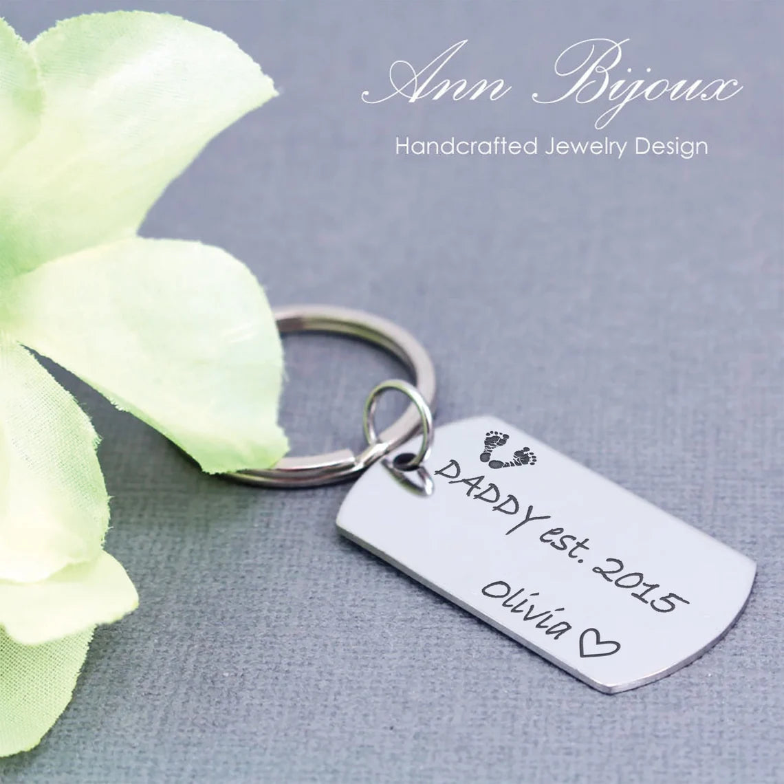 A stainless steel key chain with a laser engraved inscription 'Daddy Est' alongside a small footprint design, personalized with a name and date.