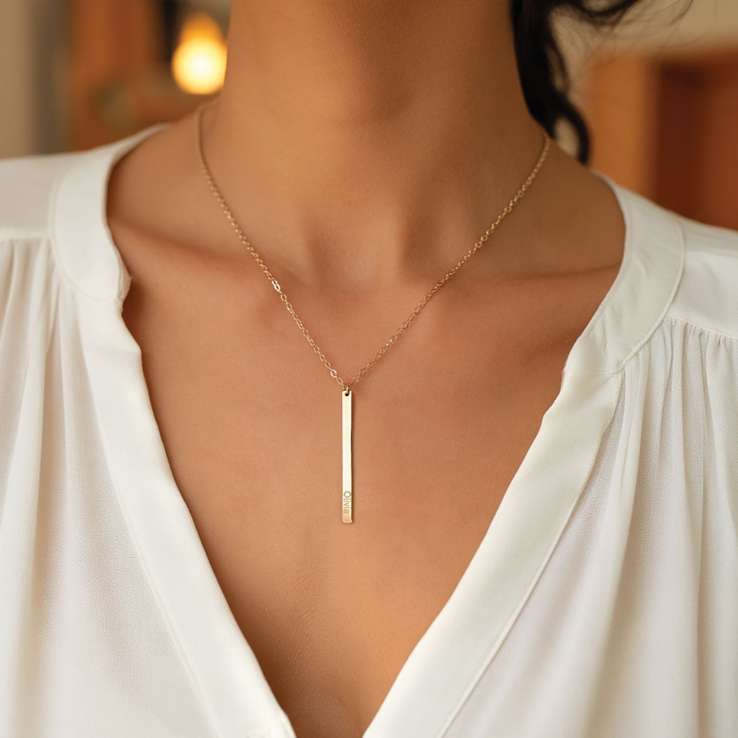 A close-up of a woman wearing a delicate gold necklace with a slim vertical bar pendant, styled with a white blouse.