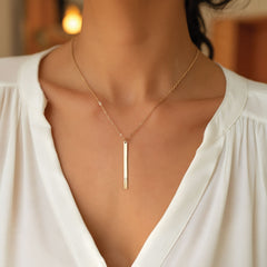 A close-up of a woman wearing a delicate gold necklace with a slim vertical bar pendant, styled with a white blouse.