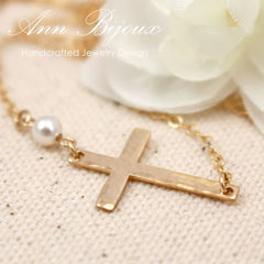 A gold-filled hammered sideway cross necklace with a flat silver cable chain and a pearl bead, displayed on a white background with a white flower.