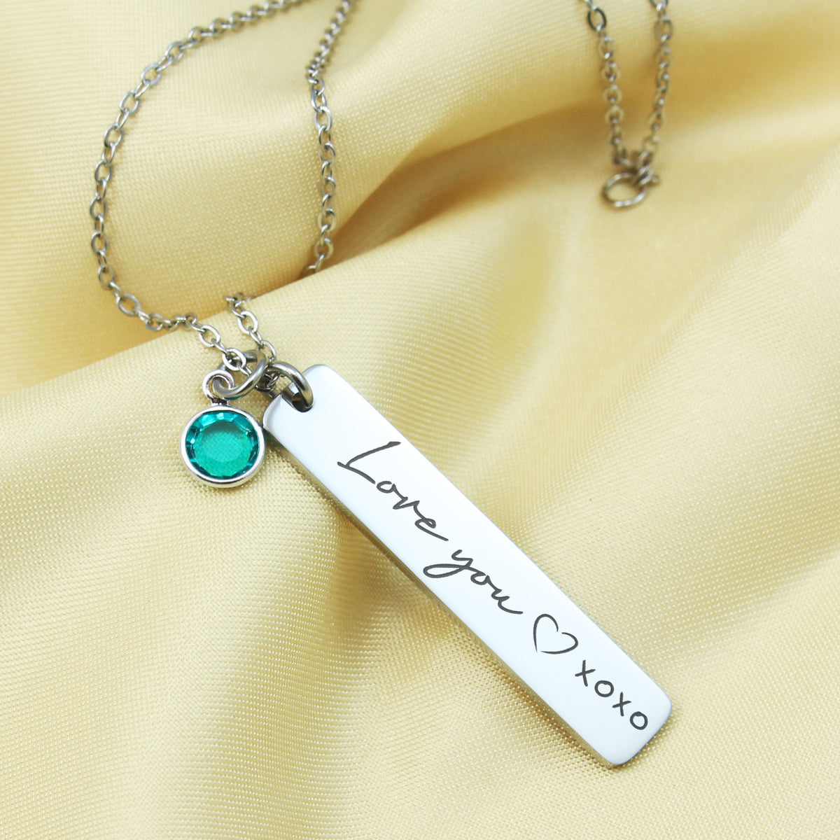A stainless steel bar necklace with a chain, featuring an engraved message 'I love you' with 'XOXO' inside a heart shape, and a small green Swarovski birthstone pendant.