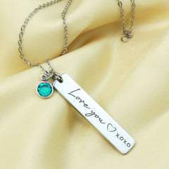 A stainless steel bar necklace with a chain, featuring an engraved message 'I love you' with 'XOXO' inside a heart shape, and a small green Swarovski birthstone pendant.