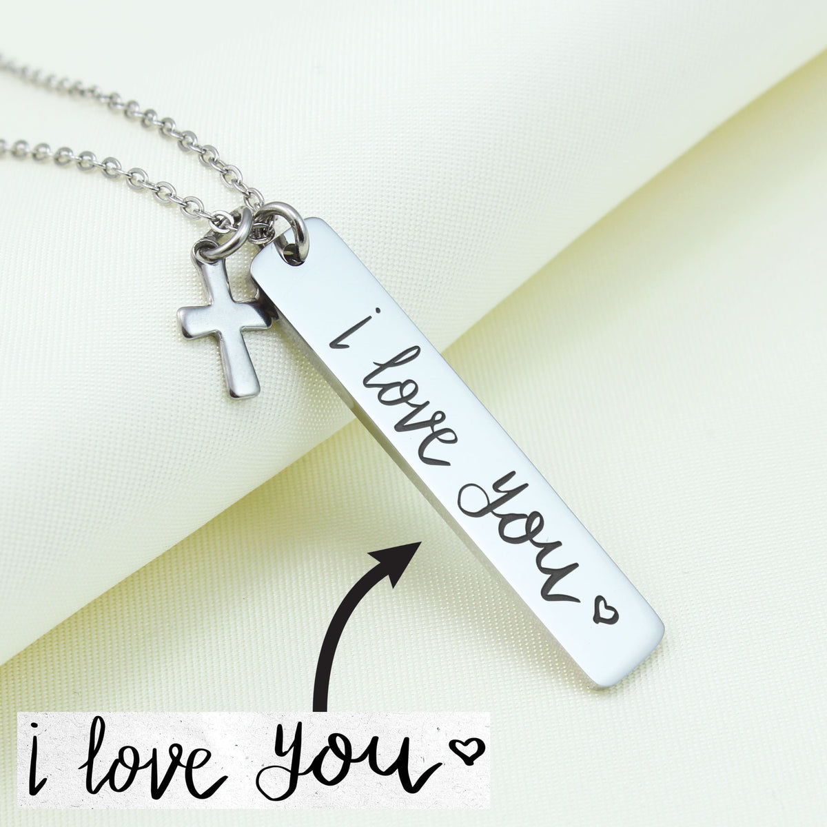 A stainless steel necklace with a cross charm and a pendant that says 'I love you' in cursive script, displayed on a white fabric background.