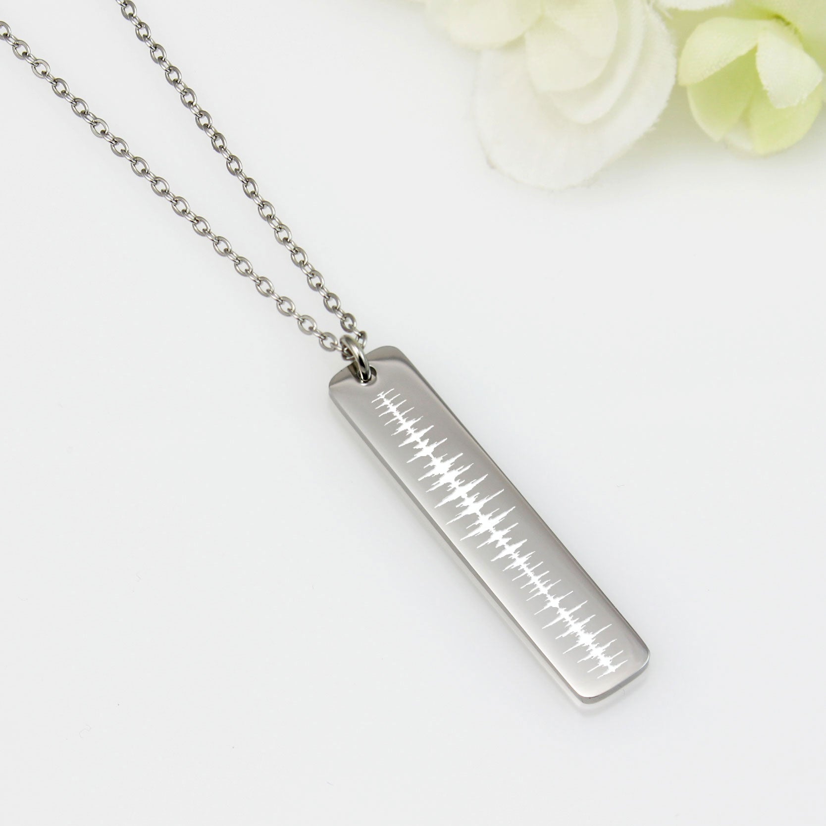 A stainless steel vertical bar pendant necklace with a chain, displayed against a white background with some floral accents.