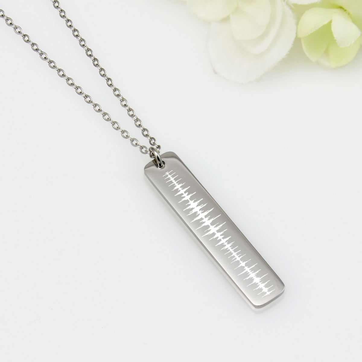 A stainless steel vertical bar pendant necklace with a chain, displayed against a white background with some floral accents.