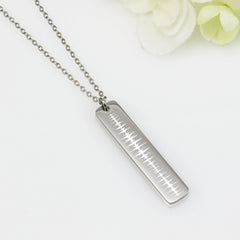 A stainless steel vertical bar pendant necklace with a chain, displayed against a white background with some floral accents.